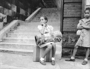 Charles "Teenie" Harris - Girl holding rolled blanket and seated on suitcase, with another girl on right, on sidewalk, ca. 1940