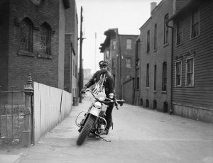 Charles "Teenie" Harris - Woman wearing dark cap with eagle, mounting motorcycle decorated with flags in an alley, ca. 1940-1946