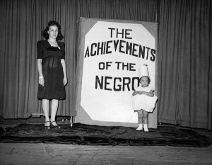 Charles "Teenie" Harris - Woman with Vickie Merritt in front of sign reading “Achievements of the Negro,” Central YWCA, Washington, Pennsylvania, February 1945